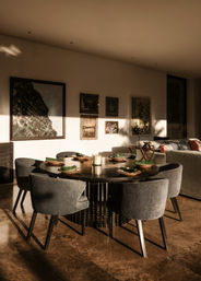 Sunlit modern dining room in a contemporary home — round dark wood table set for six with gray upholstered chairs, folded green napkins, framed wall art and a cozy living area visible in the background.