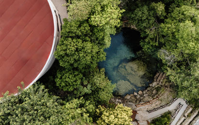 Aerial view of a turquoise cenote surrounded by lush tropical trees, rocky limestone edge, winding stone pathway and wooden railings beside a red-roofed circular building.