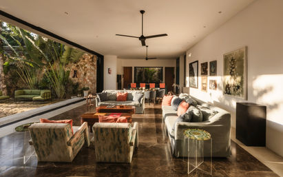 Sunlit modern open-plan living room with large sliding glass wall opening to a tropical stone patio, neutral sofas and armchairs with colorful cushions, wooden coffee tables, dining area with red chairs, ceiling fans, and lush greenery.
