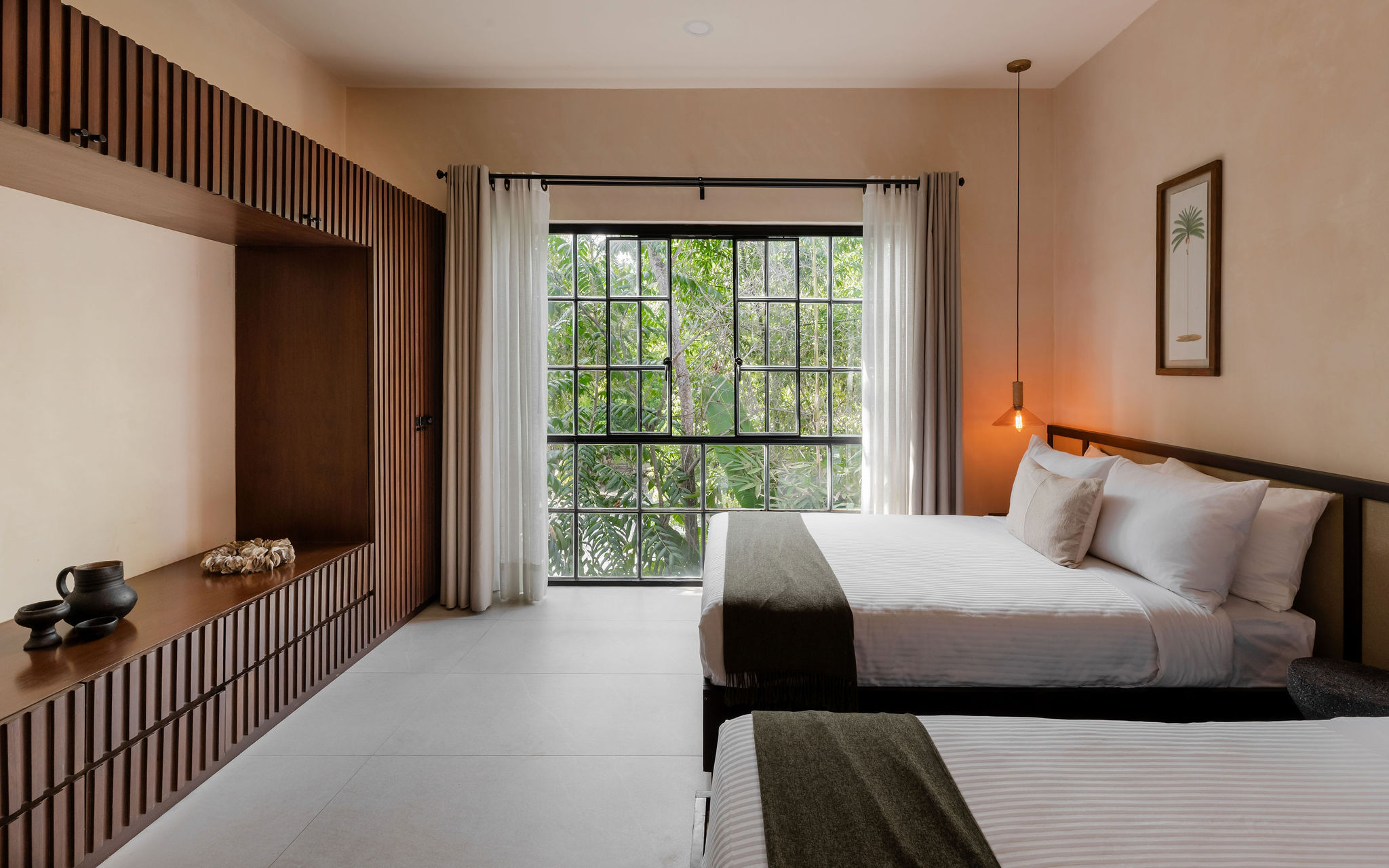 Minimalist modern hotel bedroom with two neatly made beds in white linens and olive throws, large grid window framing lush tropical greenery, wooden slatted built-in bench and warm pendant lighting