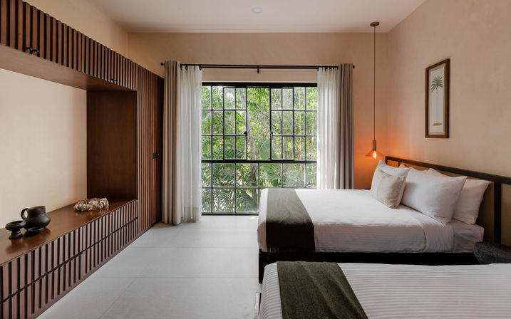 Minimalist modern hotel bedroom with two neatly made beds in white linens and olive throws, large grid window framing lush tropical greenery, wooden slatted built-in bench and warm pendant lighting