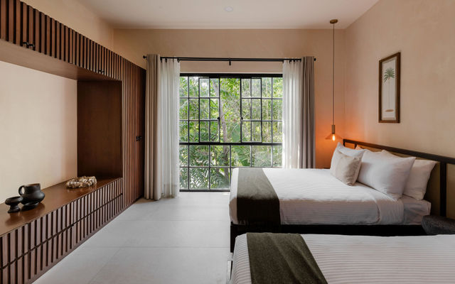 Minimalist modern hotel bedroom with two neatly made beds in white linens and olive throws, large grid window framing lush tropical greenery, wooden slatted built-in bench and warm pendant lighting