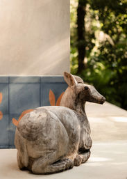 Sunlit carved wooden deer sculpture reclining on a patio next to blue decorative tiles with orange leaf accents and lush green garden foliage