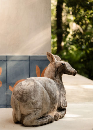 Sunlit carved wooden deer sculpture reclining on a patio next to blue decorative tiles with orange leaf accents and lush green garden foliage