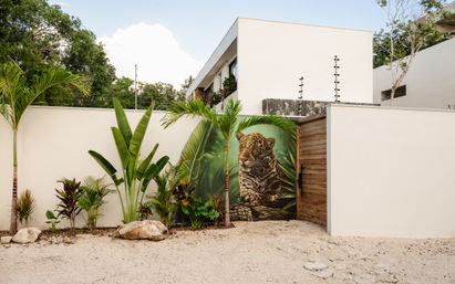 Tropical courtyard with a wooden gate featuring a bold jaguar mural, flanked by palm and banana plants against white stucco walls and a modern villa backdrop