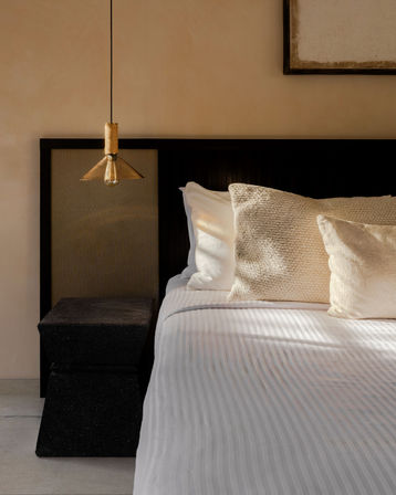 Sunlit modern bedroom close-up with white striped bedding and textured beige throw pillows against a dark wood headboard, black geometric stone nightstand and hanging brass pendant light on a warm beige wall.