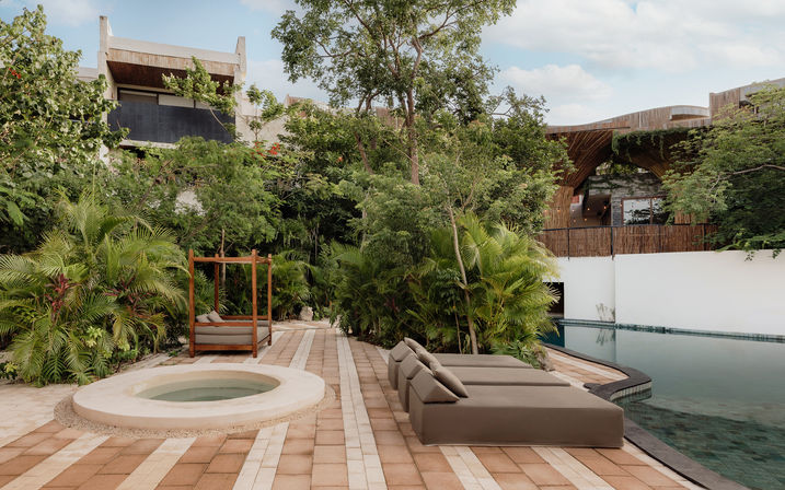 Tropical resort pool deck with round hot tub, three cushioned sun loungers, a wooden canopy daybed and lush greenery framing modern wood-and-stone architecture.