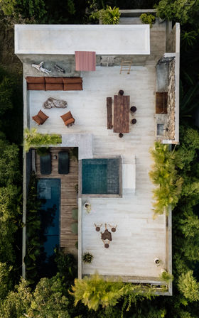 Aerial drone view of a tropical villa rooftop terrace with outdoor dining table, cushioned lounge seating, a square plunge pool and adjacent lap pool, sun loungers on a wooden deck, and lush green foliage surrounding the space.