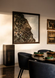 Sunlit modern dining room with round dark-wood table set with green napkins, navy upholstered chairs, and a large framed abstract cityscape painting on the wall.