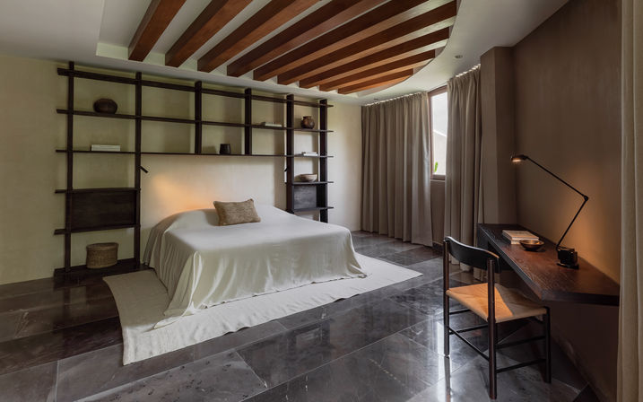 Minimalist modern bedroom with low white-linen bed, dark wood shelving and ceiling beams, glossy stone floor, desk and lamp by a curtained window in neutral tones.