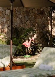 Pink plastic flamingo lawn ornament among palm plants and a stone wall on a sunlit tropical poolside patio with lounge chairs.