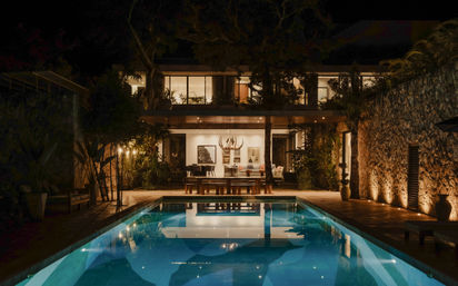 Nighttime view of a modern two-story villa with warm interior lights, outdoor dining area and a glowing rectangular swimming pool reflecting chandeliers, stone walls and tropical plants.