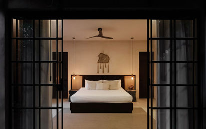 Modern minimalist bedroom seen through black-framed sliding glass doors — king bed with white linens, pendant bedside lights, ceiling fan and woven wall hanging in a cozy resort-style villa interior.