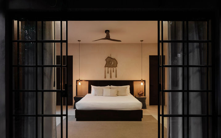 Modern minimalist bedroom seen through black-framed sliding glass doors — king bed with white linens, pendant bedside lights, ceiling fan and woven wall hanging in a cozy resort-style villa interior.