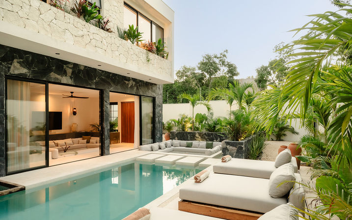 Modern tropical villa patio with turquoise plunge pool, cushioned sunbeds and built-in sofa, sliding glass doors to a living room, lush palm landscaping and stone accents