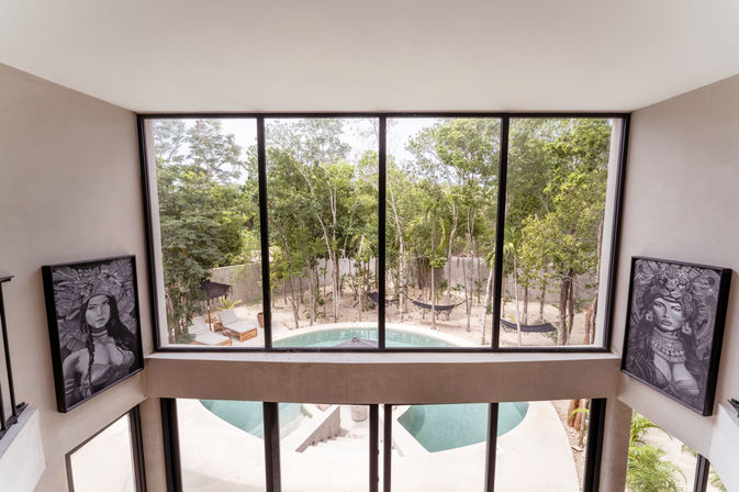 View through black-framed floor-to-ceiling windows showing a curved pool, lounge chairs and hammocks in a leafy tropical courtyard, flanked by two large monochrome portrait prints.