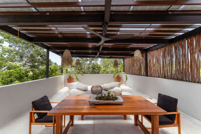 Covered tropical outdoor terrace with dark wooden pergola and ceiling fans, rattan pendant lights, a wooden dining table with black cushioned chairs, low white floor cushions, potted plants and lush green foliage view.