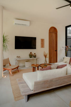 Modern boho living room in neutral tones with a low wooden sofa and cream cushions, round wooden nesting tables, wall-mounted TV, tall potted plant, arched wooden doorway and woven jute rug.