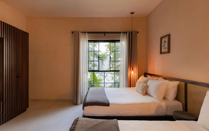 Cozy modern bedroom with two neatly made beds, warm beige walls, wooden headboards and slatted wardrobe, sheer curtains framing a large window with lush tropical greenery outside, and a glowing pendant light for ambiance