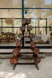 Rustic wooden dumbbells marked 10–60 lb stacked on a metal rack on a gravel patio in front of a modern glass-walled home, tropical plants, dining table and cozy lounge visible through the windows.