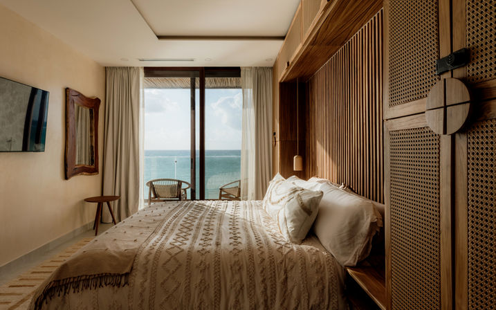 Sunlit ocean-view bedroom in a coastal suite with wooden slatted walls, textured bedding, and sliding glass doors opening to a balcony with wicker chairs and turquoise sea.