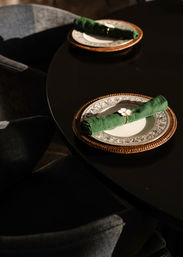 Sunlit elegant dining table setting with decorative china on a woven rattan charger and a rolled green linen napkin held by a small flower napkin ring on a dark glossy table.
