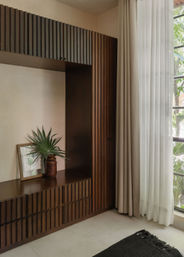 Sunlit modern interior corner with dark wood slatted built-in cabinet, white sheer curtains, potted palm-leaf in a ceramic vase, and tiled floor beside a window with garden greenery.