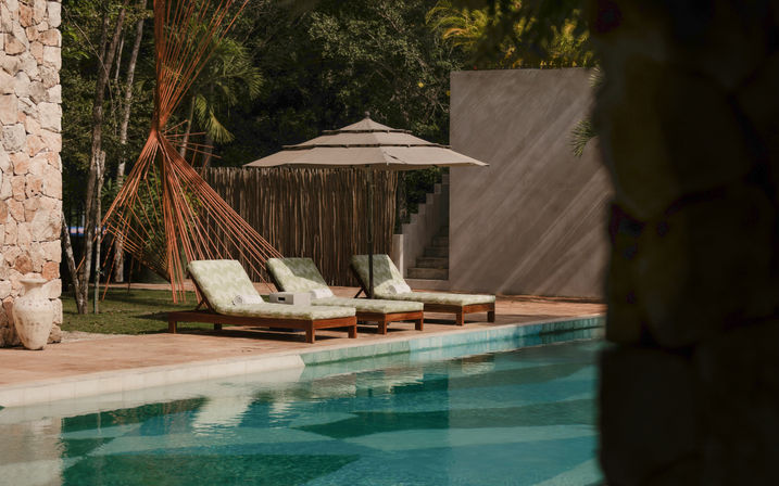 Three cushioned wooden lounge chairs under a beige umbrella beside a turquoise pool, with a stone wall, palms and sunlit patio at a tranquil tropical retreat.