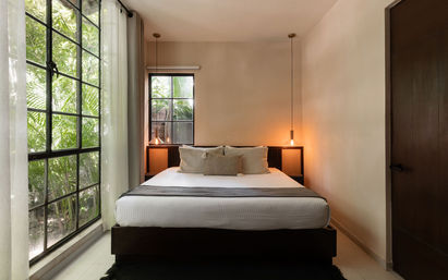 Minimalist modern bedroom with white linens, hanging pendant bedside lights, and a large black-framed window overlooking lush tropical plants and soft natural light.