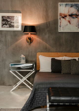 Cozy modern industrial bedroom with concrete textured wall, wooden headboard, dark quilted bedding and patterned pillows, mirrored X-leg nightstand with decorative tissue box, warm black wall sconce and framed art, and a black leather bench at the foot of the bed.