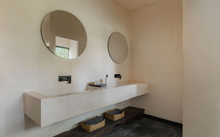 Minimalist modern bathroom featuring a long floating concrete trough sink, two round mirrors, matte-black wall faucets, neutral plaster walls and woven storage baskets on a dark tiled floor.