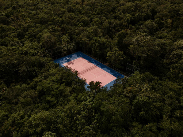 Aerial view of a pink-and-blue tennis court tucked into dense green forest, sunlight and shadows on the empty court.