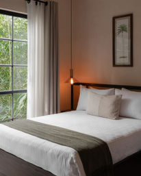 Cozy minimalist bedroom with white striped bedding, olive-gray throw and lumbar pillow, black-framed window showing lush greenery, sheer curtains, warm hanging Edison pendant and framed palm print.