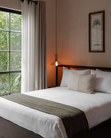 Cozy minimalist bedroom with white striped bedding, olive-gray throw and lumbar pillow, black-framed window showing lush greenery, sheer curtains, warm hanging Edison pendant and framed palm print.