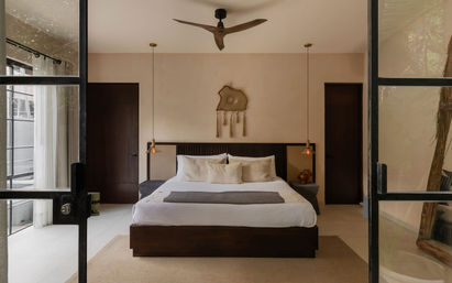 Modern minimalist bedroom with a king bed dressed in neutral linens, textured wall hanging above the headboard, twin pendant lights, wooden ceiling fan, and floor-to-ceiling glass doors opening to a balcony.