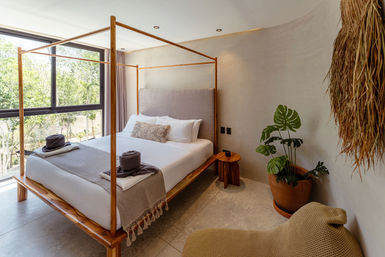 Tropical-inspired bedroom with natural wood four-poster bed, neutral linen bedding and decorative pillow, folded towels and sun hat on the foot of the bed, floor-to-ceiling window overlooking lush greenery, potted monstera and woven pendant light.
