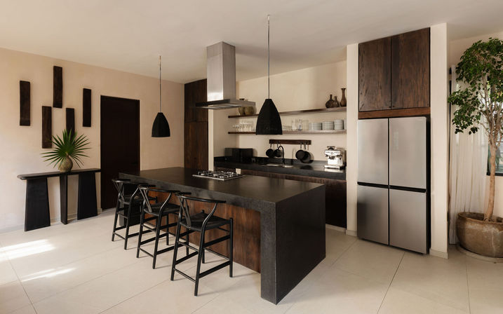 Modern open-plan kitchen with a large black stone island and four black bar stools, dark wood cabinets, stainless steel refrigerator, pendant lights, open shelves with dishes and espresso machine, and a potted plant.
