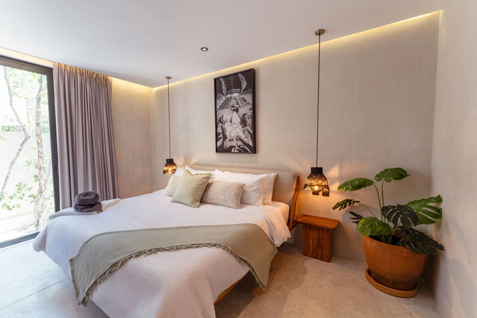 Sunlit modern minimalist bedroom with a cozy king bed, layered neutral pillows and throw, two woven pendant lamps, framed wall art, large potted monstera, and floor-to-ceiling window with curtains showing trees outside.