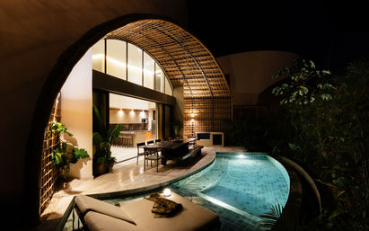 Night view of a luxury tropical villa with arched bamboo patio, open-plan kitchen, illuminated private pool, outdoor dining area and lounge chair