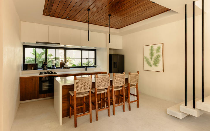 Bright modern minimalist kitchen with wood-paneled ceiling, white island and four woven-back wooden bar stools, black pendant lights and large paned window overlooking lush greenery.