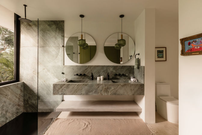 Spa-like modern bathroom with marble double-sink vanity, two round mirrors, green pendant lights, glass walk-in shower and large window with tropical plants