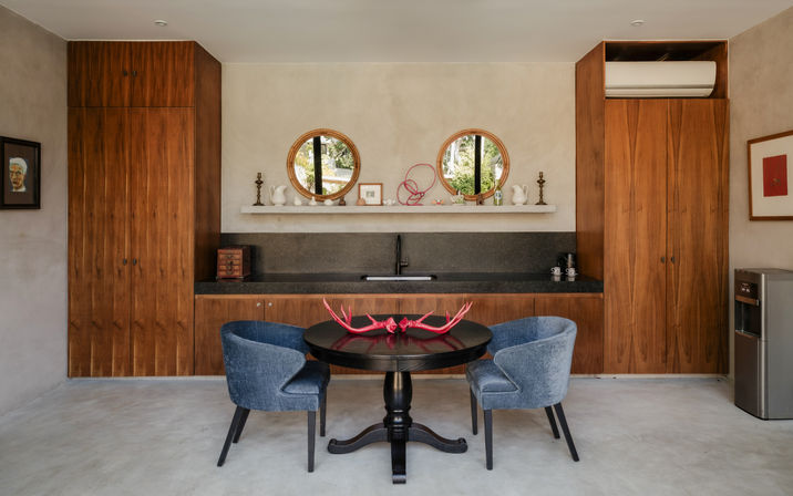 Modern kitchen dining nook with round black table and two blue upholstered chairs, wood-paneled cabinets and black stone countertop with sink, twin round mirrors and decorative shelf, playful red antler centerpiece on concrete floor.