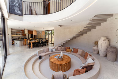 Cozy sunken circular lounge with built-in curved sofa and wooden coffee table in a modern open-plan Mediterranean home; floating concrete staircase, dining table with woven pendant lights, neutral plaster walls and large decorative urns.