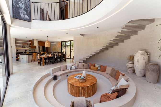 Cozy sunken circular lounge with built-in curved sofa and wooden coffee table in a modern open-plan Mediterranean home; floating concrete staircase, dining table with woven pendant lights, neutral plaster walls and large decorative urns.