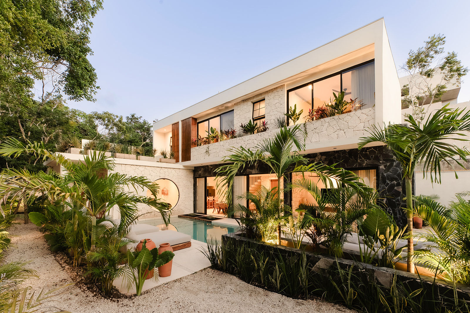 Two-story modern tropical villa with stone facade and floor-to-ceiling windows, sunset-lit interiors, private outdoor pool, lush palm landscaping and cozy lounge area
