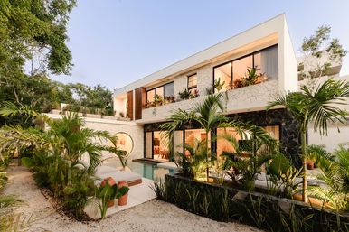 Two-story modern tropical villa with stone facade and floor-to-ceiling windows, sunset-lit interiors, private outdoor pool, lush palm landscaping and cozy lounge area
