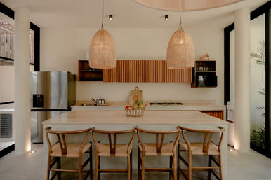 Bright open-concept modern kitchen with a large island, four woven-seat wooden bar stools, oversized rattan pendant lights, slatted wood cabinets, stainless refrigerator and a fruit basket centerpiece.