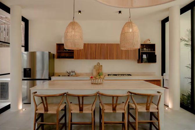 Bright open-concept modern kitchen with a large island, four woven-seat wooden bar stools, oversized rattan pendant lights, slatted wood cabinets, stainless refrigerator and a fruit basket centerpiece.