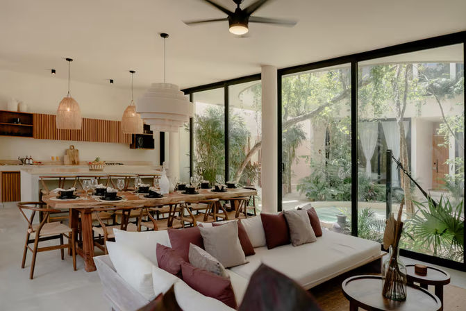 Sunlit open-plan modern villa interior with indoor-outdoor living: wooden dining table set with place settings, woven pendant lights, white sectional sofa with burgundy and gray pillows, and floor-to-ceiling glass doors opening to a lush tropical courtyard and small pool.