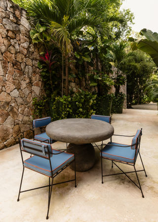 Sunlit tropical patio courtyard with a round concrete pedestal table and four blue-cushioned metal chairs against a rustic stone wall, surrounded by palms and lush greenery.